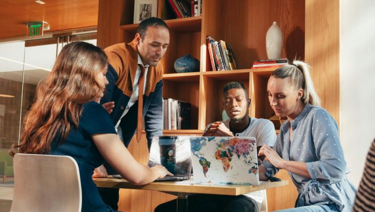 Four students sit studying around a table in a bright, modern university space. Two open laptops are on the table, behind them is a wooden bookshelf filled with books and decorative items. | links to article: Disabled Students’ Allowance: Our Policy Work and Impact