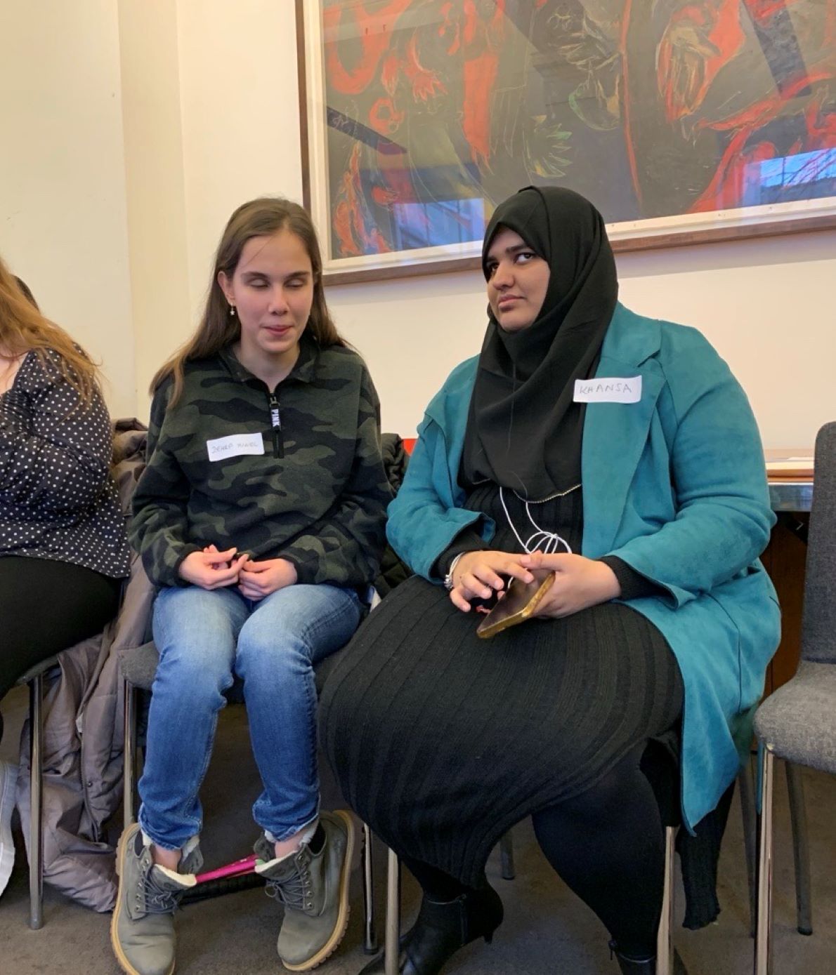 Khansa is sitting on a chair with her hands on her lap, talking to a delegate next to hear, wearing all black with a blue coat.