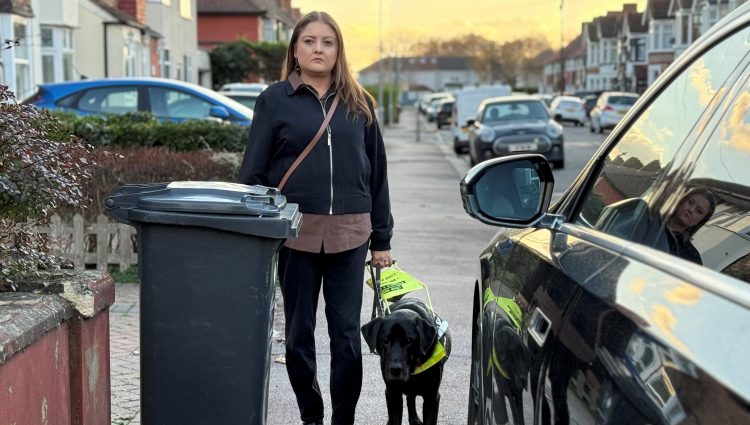 Bhavini Makwana, a South Asian woman, stopped on a pavement with her Guide Dog who has its hind legs on the road. Bhavini is blocked from walking further because a car is parked on the pavement. The guide dog appears ready to lead her out on to the road.