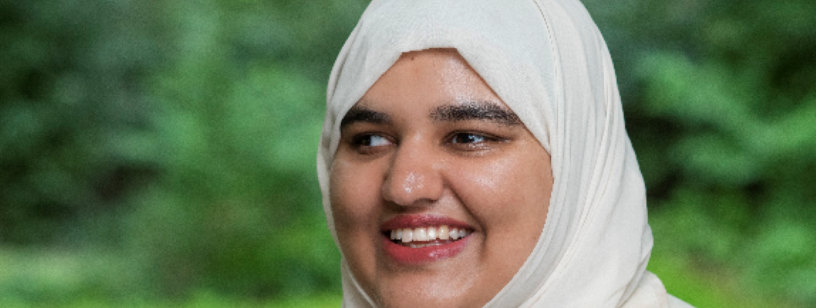 A picture of Khansa smiling at the camera, wearing a light coloured hijab and matching coat, standing in a green outdoor space.