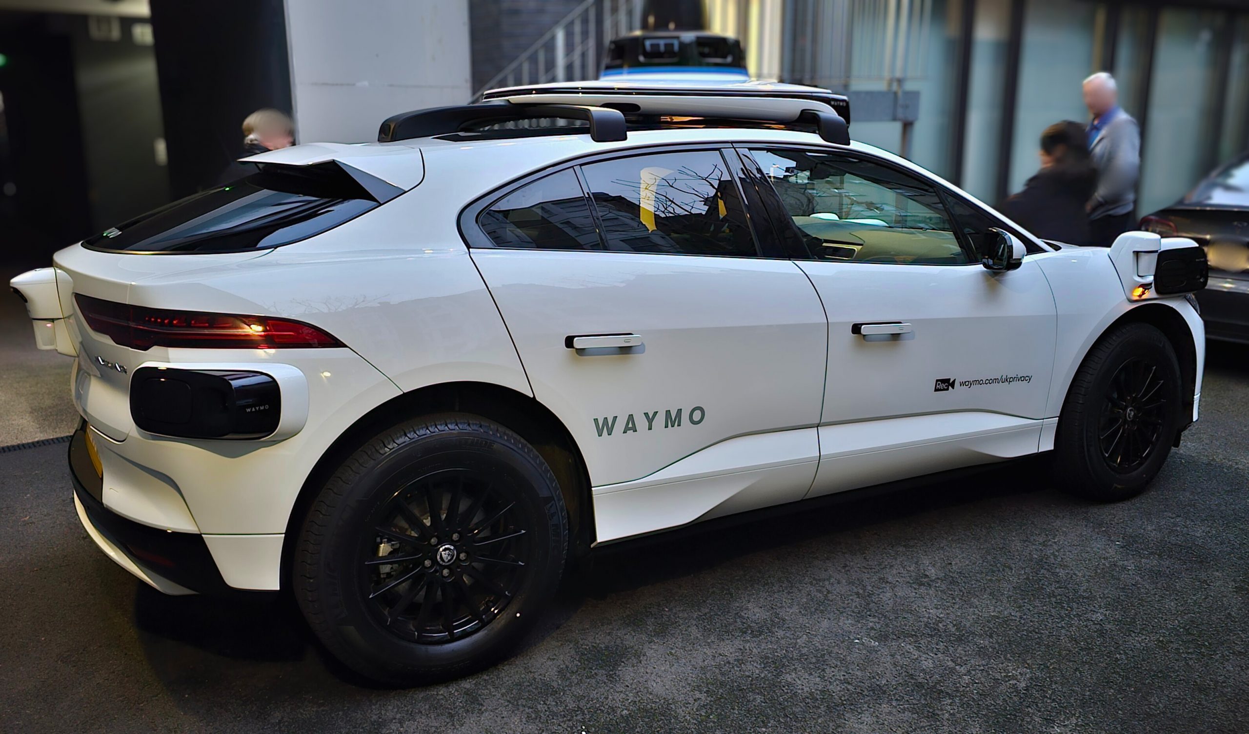 Side view of a white, electric Waymo vehicle featuring black wheels and rooftop lidar equipment.