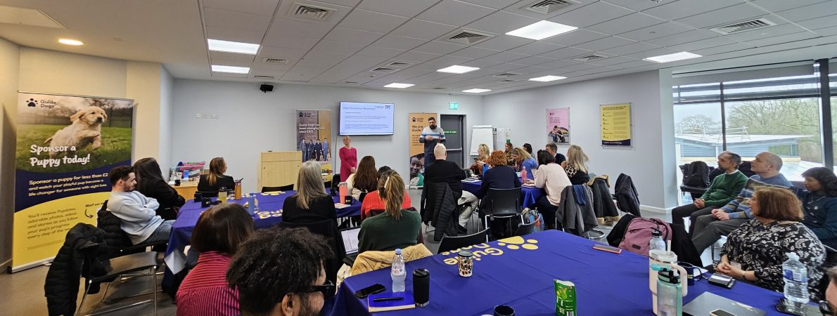 Large group of people seated around tables in a bright training room, listening to Jack and Kelly, TPT presenting, standing at the front near a screen. The tables are covered in blue cloths with Guide Dogs' logo, and posters about guide dogs and puppy sponsorship are displayed around the room.