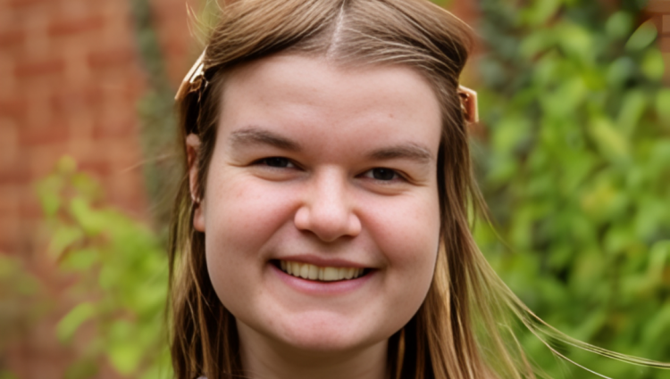 Close up of Sarah, a woman in her early twenties with shoulder length light brown hair. Sarah smiles at the camera outdoors in front of a blurred background of summer foliage.