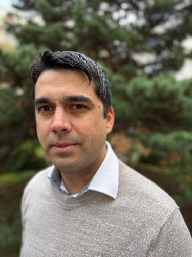 A headshot-style photo of Gareth, a man with short black hair, smiling. He can be seen wearing a white collared shirt with a cream long-sleeved jumper over the top. The background is a blurred outdoor scene with dark greenery.