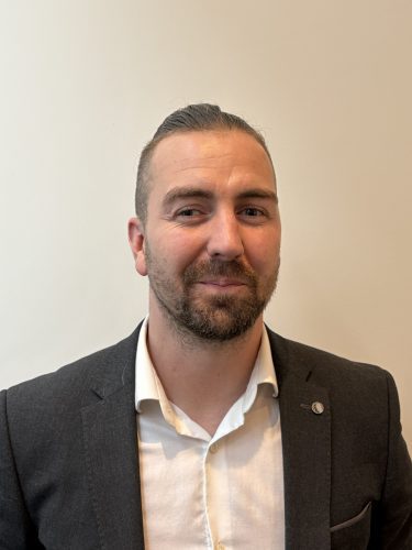 A headshot style photo of Josh, a man with dark hair done up into a Top Knot with a short dark beard. Josh can be seen wearing a white button-up shirt with a smart dark blazer jacket. He is stood in front of a white wall with warm lighting.