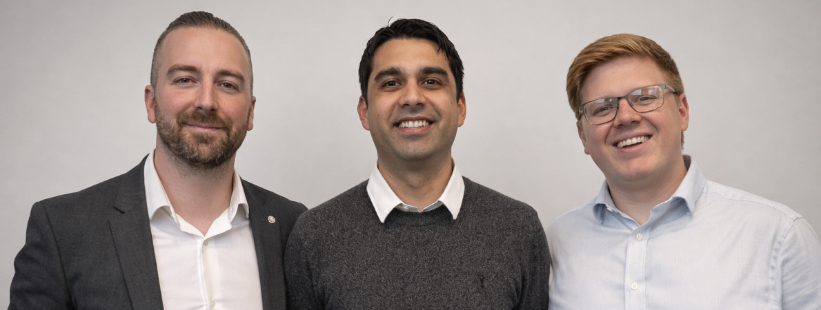 This image shows a group photo of the GSP team, from left to right, we have: Joshua Feehan (Head of Internships), Gareth Dawson (Internship Coordinator), Charlie Rashbrook (Internship Coordinator). All three are stood against a white wall with warm lighting. The image is taken from the sternum height. Joshua - a man with dark hair done up into a Top Knot with a short dark beard. Josh can be seen wearing a white button-up shirt with a smart dark blazer jacket. Gareth - , a man with short black hair, is smiling. He can be seen wearing a white collared shirt with a dark grey woollen long-sleeved jumper over the top Charlie - a young man with ginger hair swept to the right. He is bespectacled and wearing a light green button shirt with a collar. Charlie is smiling.