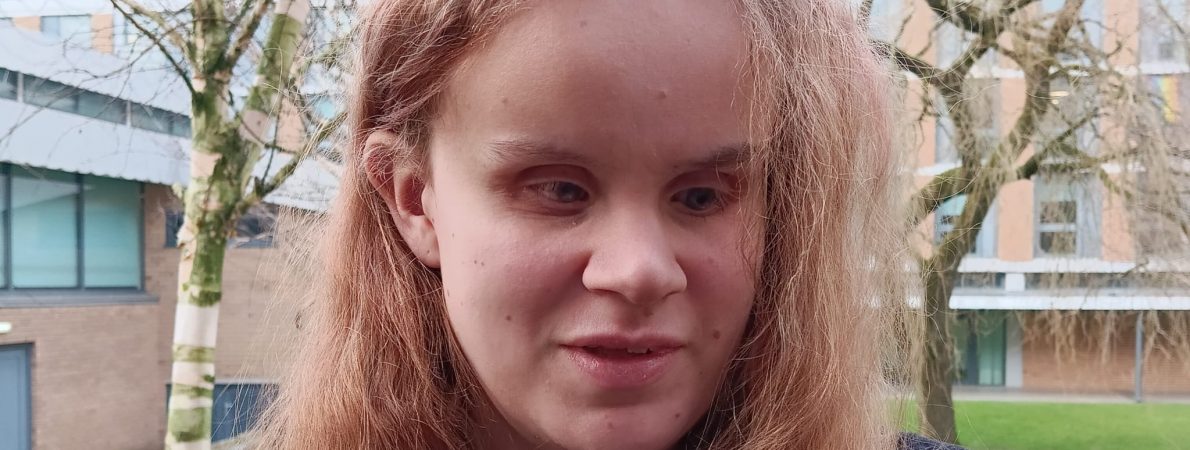 A picture of Martha standing outside in a grassy area with a leafless trees and brick buildings, wearing a dark coat and a purple top.
