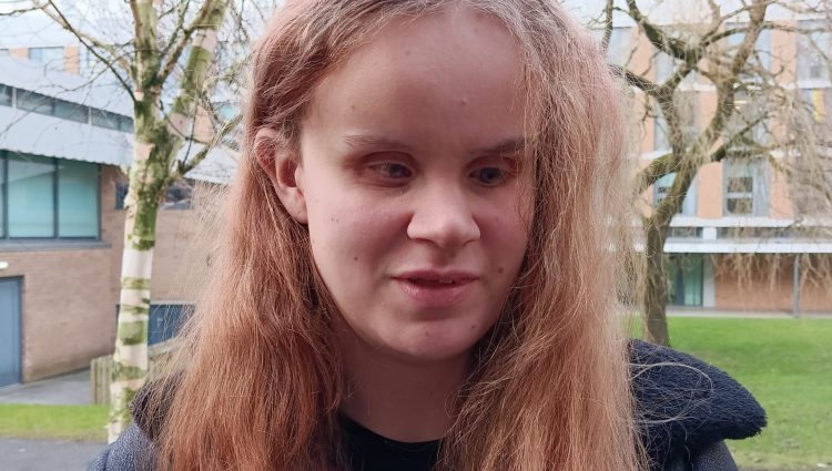 A picture of Martha standing outside in a grassy area with a leafless trees and brick buildings, wearing a dark coat and a purple top. | links to article: Fighting For Access to University