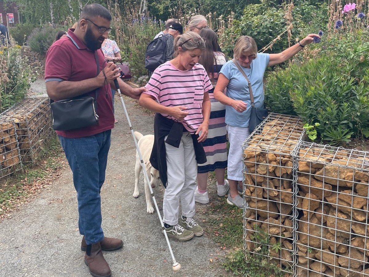 Bedfordshire SLC members are walking together along a gravel garden path bordered by raised stone-filled wire planters. One member is at the front is using a long white mobility cane to feel the ground ahead, while another nearby, holds a guide dog on a harness. The garden is lush with a variety of green plants, tall flowers, and purple blooms in the background. One person reaches out to touch a plant growing from the raised planter.
