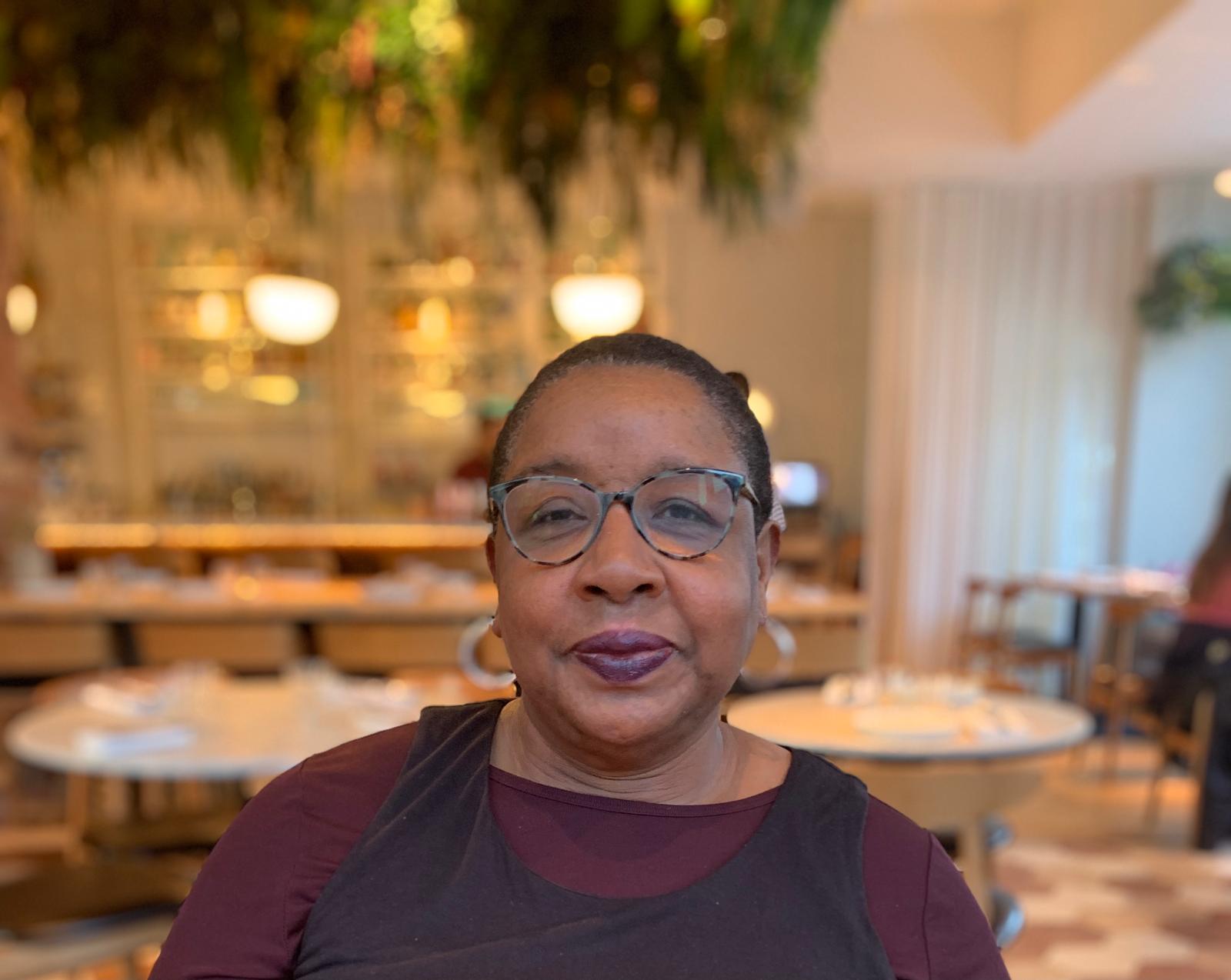 Photo of TPT’s Beverley Duguid, smiling, wearing glasses, with short black hair. Restaurant tables and chairs are in the background.