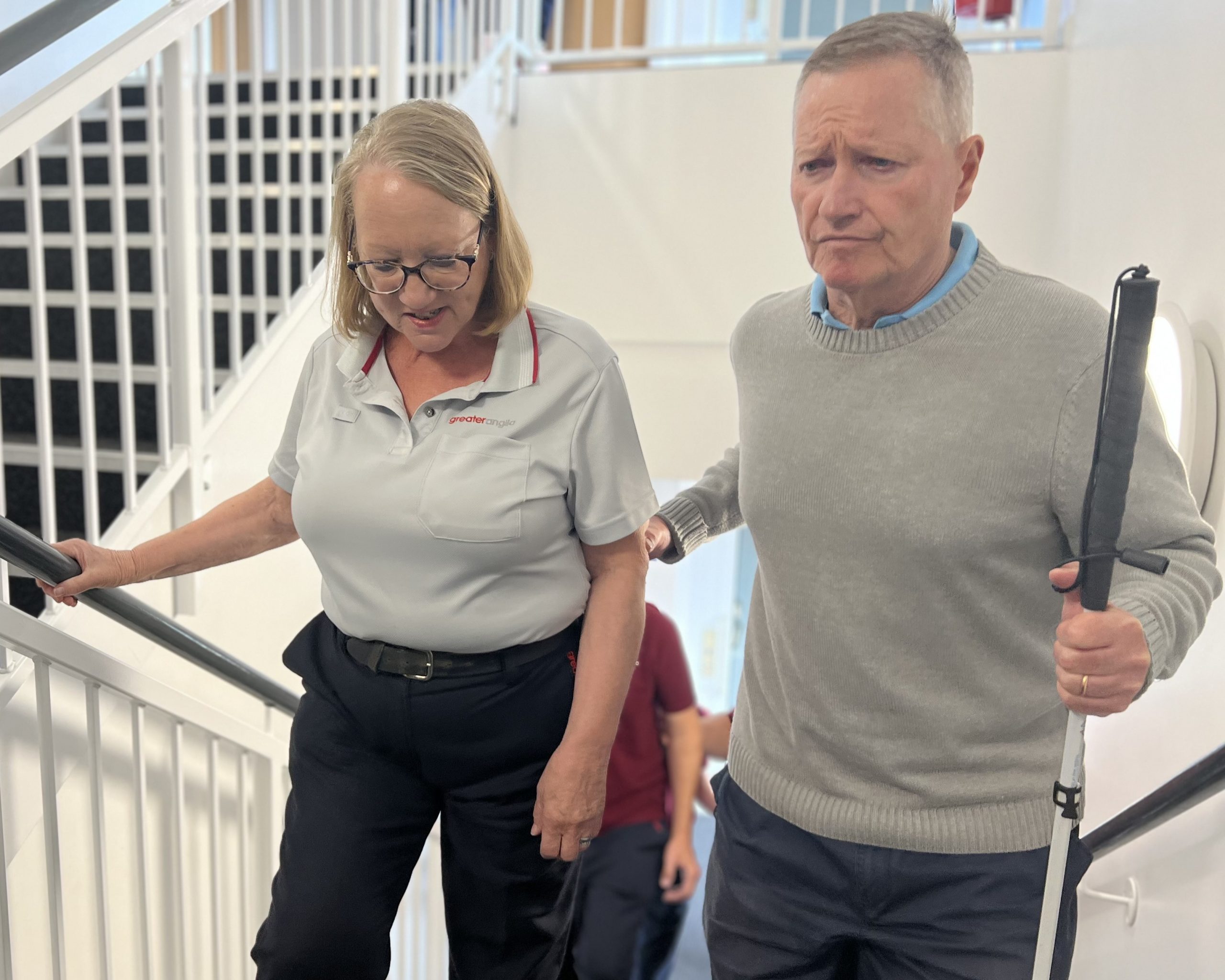 A female Greater Anglia staff member guides Harry, Essex SLC member, up the stairs during the sighted guiding element of the VI Awareness Session. Harry is holding onto the lady's arm with one hand, and his long cane in the other.