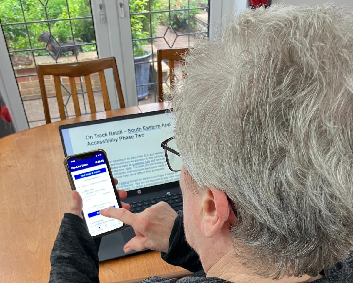 Leslie, North Yorkshire SLC member, is pictured at her table, holding a smartphone. The smartphone displays a rail ticket booking app with a prominent “Find times & tickets” button. In front of the person is an open laptop showing a document titled “On Track Retail – South Eastern App Accessibility Phase Two.