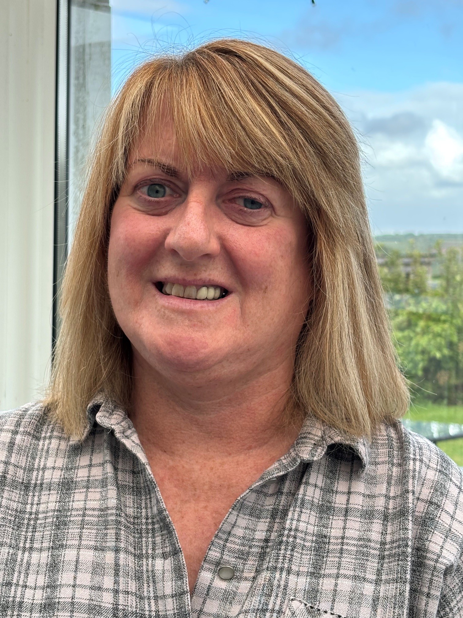 TPT’s Maria Moorhouse smiling with shoulder length blond hair wearing a checkered top. She is stood indoors infront of a window with blue sky and greenery in the background.