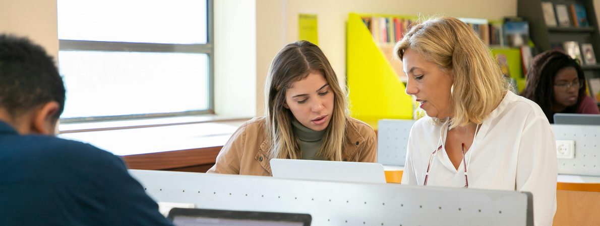 A picture of two women working at a desk in a bright library, focusing on a laptop in front of them.