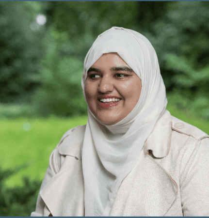 Khansa, Student Voices member, looks to her side. She wears a light hijab and beige coat and sits outdoors. Green foliage softly blurs in the background.