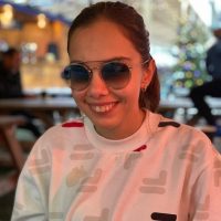 Image shows Zehra, Student Voices member, smiling at the camera. Her hair is pulled back in a ponytail, she is wearing a white jumper, and sunglasses.