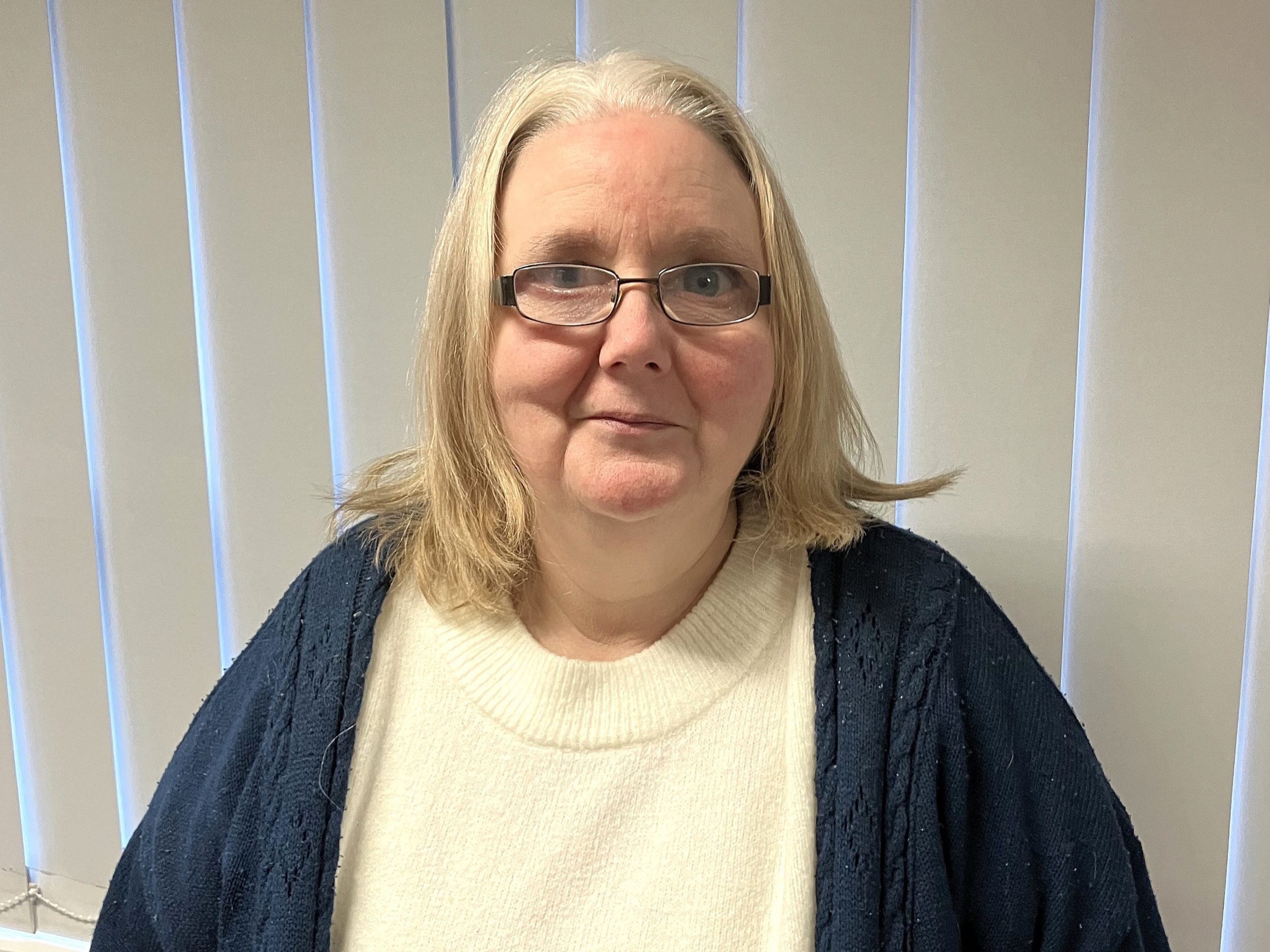 Photo of Tanja, Staffordshire Sight Loss Council member. Tanja has shoulder-length, fair hair and is wearing dark rimmed glasses. She is wearing a cream knitted top beneath a dark cardigan and is smiling at the camera.