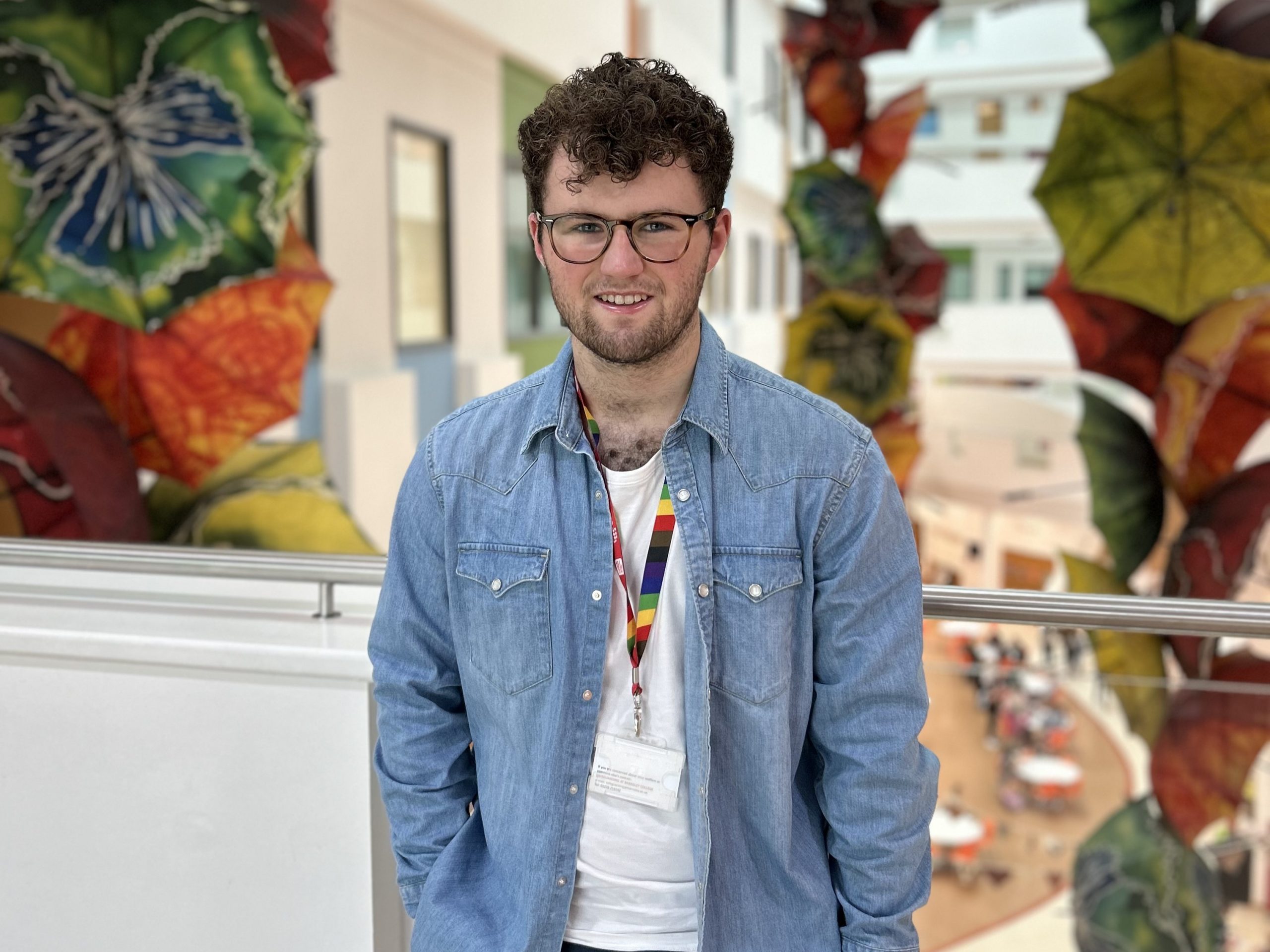 Tom, Young Voices member wearing glasses and a denim shirt stands indoors with a rainbow lanyard, with large colourful hanging decorations and an open atrium in the background.