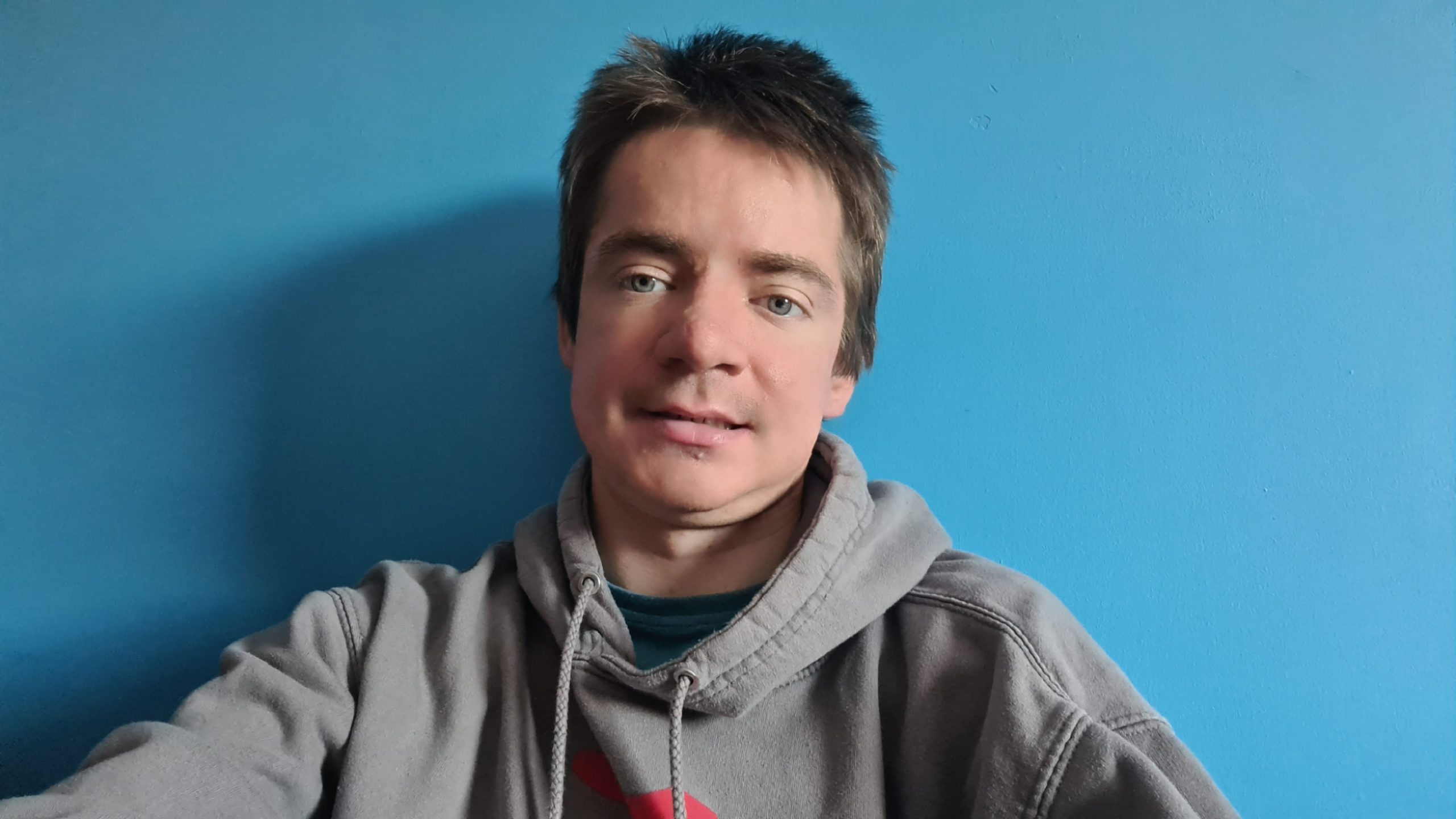 Close-up portrait of Ben Wright with short hair wearing a grey hoodie, sitting against a plain blue wall and facing the camera. 