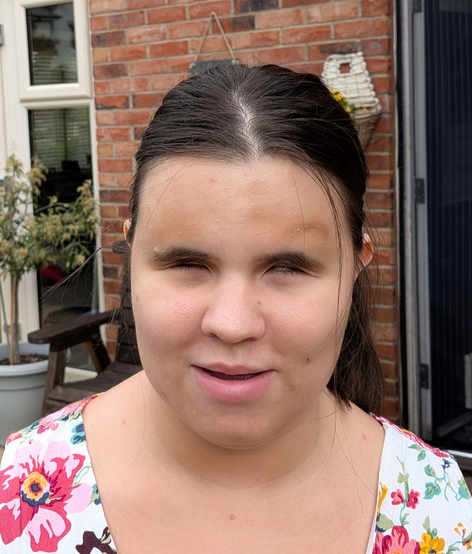 Close-up jheadshot of a Carys standing outdoors in front of a brick house, with a window, patio chair and hanging basket visible in the background.