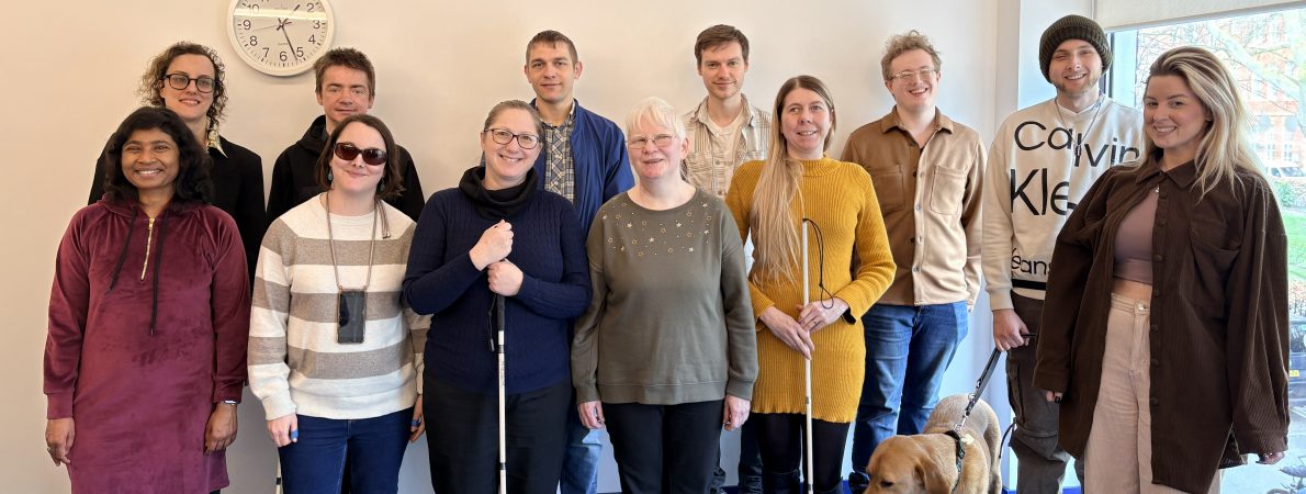 Group shot of 12 male and female interns from the 2025-26 cohort stood at the back of an office room with a blue carpet and cream walls. A guide dog can be seen in the photo. Several of the interns are holding a white navigation cane. They are smiling and look happy.