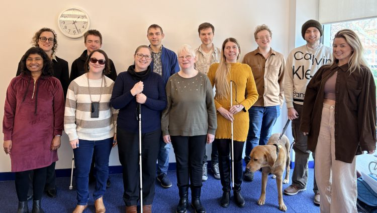 Group shot of 12 male and female interns from the 2025-26 cohort stood at the back of an office room with a blue carpet and cream walls. A guide dog can be seen in the photo. Several of the interns are holding a white navigation cane. They are smiling and look happy.