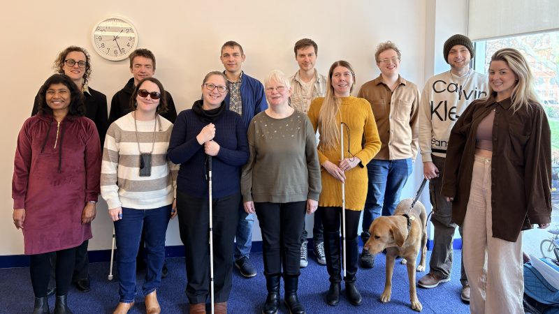 Group shot of 12 male and female interns from the 2025-26 cohort stood at the back of an office room with a blue carpet and cream walls. A guide dog can be seen in the photo. Several of the interns are holding a white navigation cane. They are smiling and look happy.