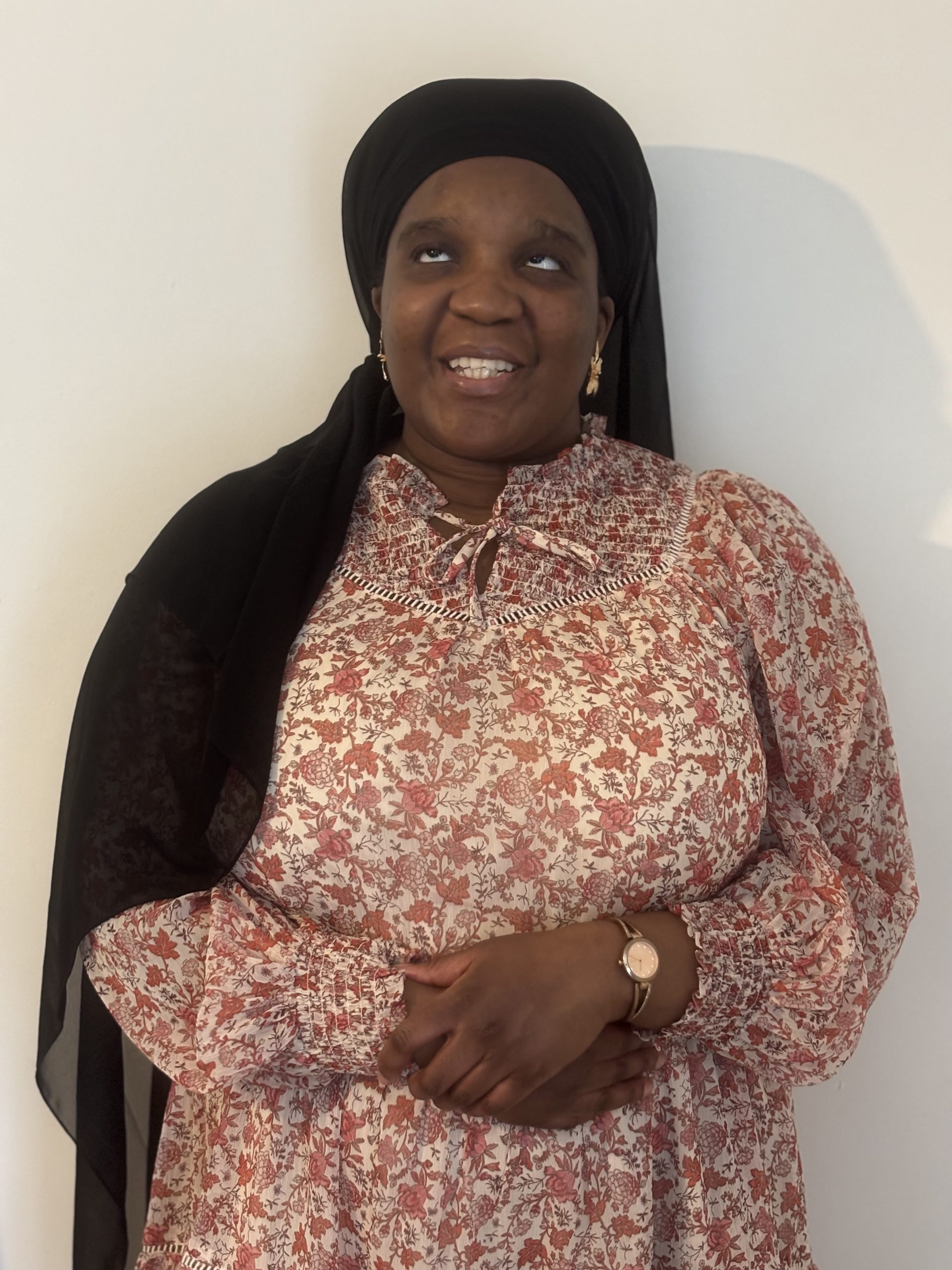 Laura standing indoors against a plain wall, wearing a patterned dress and a dark headscarf, and a petite gold watch and earrings.