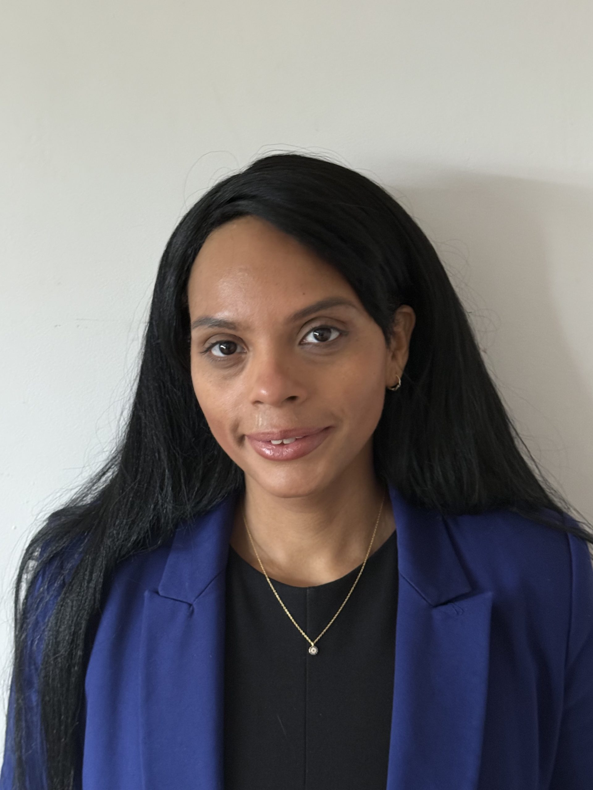 Head-and-shoulders portrait of Yarissa with long dark hair, wearing a blue blazer over a dark top and a small pendant necklace, standing against a plain light-coloured wall. 