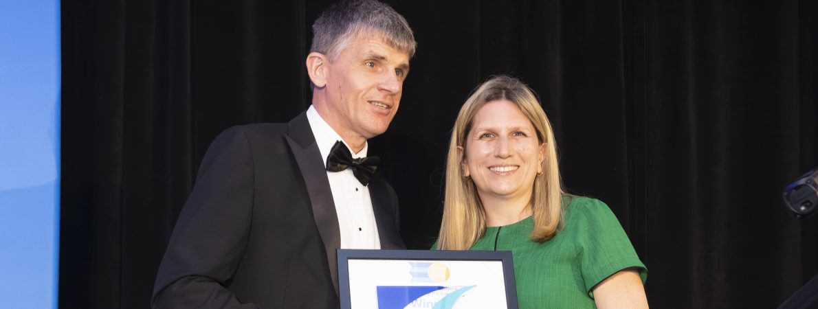 Dave Smith, Engagement Manager for South East England, stands on stage with a CIHT Awards presenter. Dave is wearing a black tuxedo and holding a long cane, while the presenter is wearing a green dress. A dark curtain forms the backdrop, with stage lighting visible to one side. Dave and the presenter are holding a framed award and trophy for the camera.