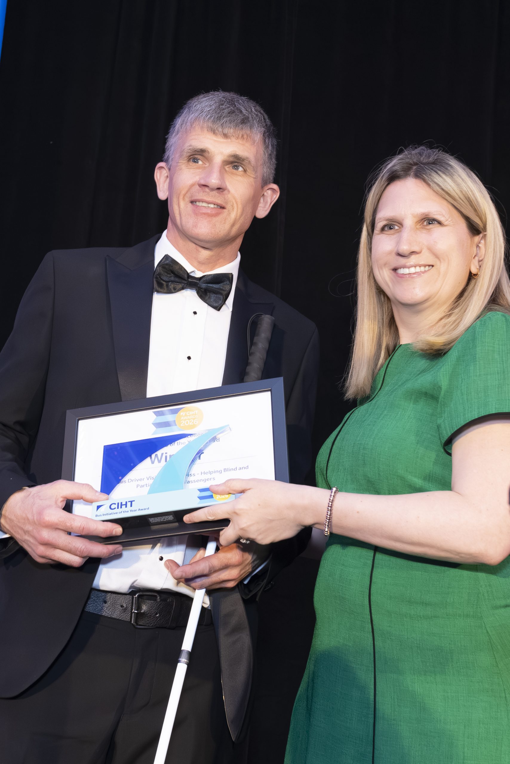Dave Smith, Engagement Manager for South East England, stands on stage with a CIHT Awards presenter. Dave is wearing a black tuxedo and holding a long cane, while the presenter is wearing a green dress. A dark curtain forms the backdrop, with stage lighting visible to one side. Dave and the presenter are holding a framed award and trophy and smiling for the camera.