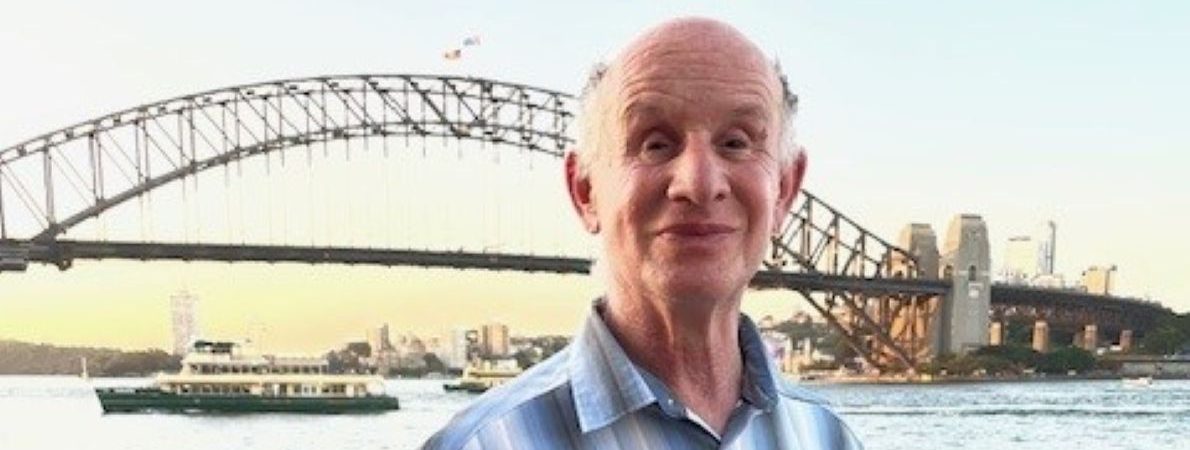 Jeff stands in front of a bridge crossing a wide river during a sunset. A city skyline can be seen on the other side of the bridge. Jeff is dressed for sunny weather and smiling at the camera.