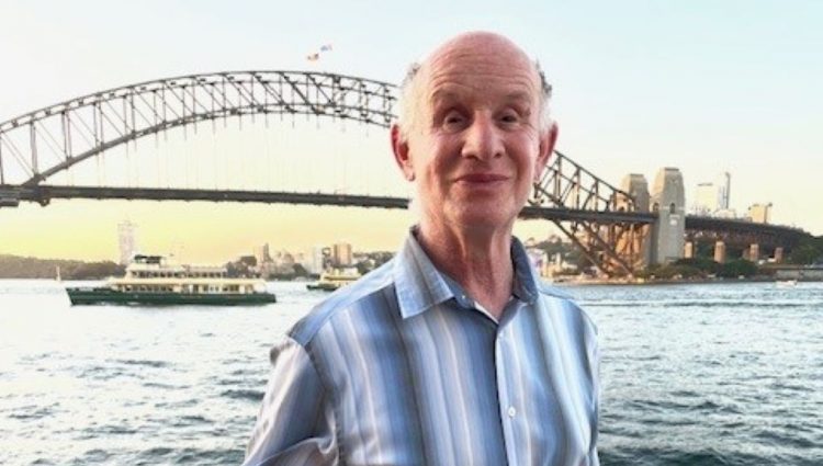 Jeff stands in front of a bridge crossing a wide river during a sunset. A city skyline can be seen on the other side of the bridge. Jeff is dressed for sunny weather and smiling at the camera. | links to article: Supporting Blind and Partially Sighted Adult Learners: Jeff’s Story
