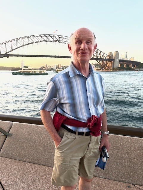 Jeff stands in front of a bridge crossing a wide river during a sunset. A city skyline can be seen on the other side of the bridge. Jeff is dressed for sunny weather and smiling at the camera.