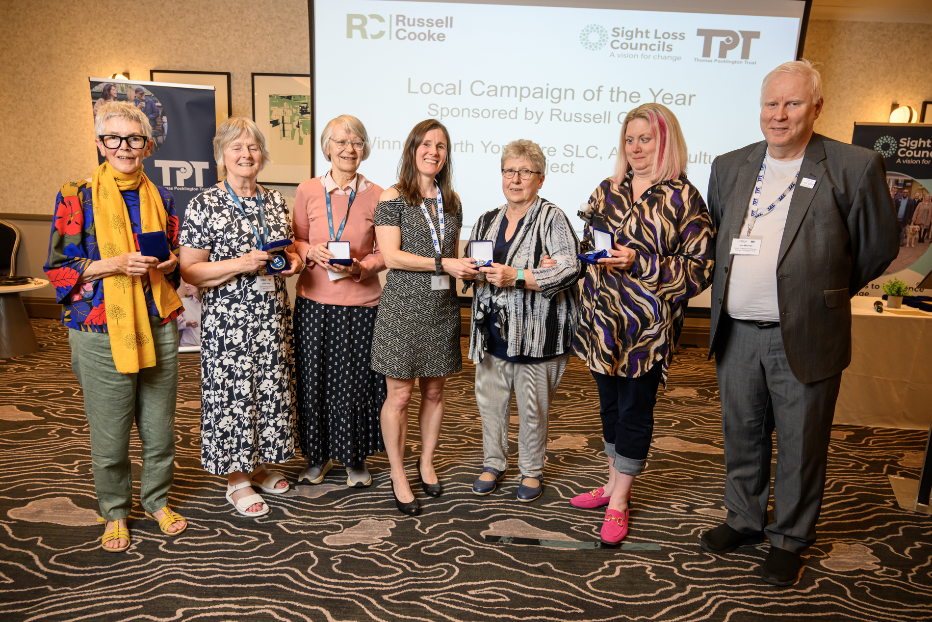 Members of North Yorkshire SLC stand together at the front of the conference room, face the camera, holding their winners medals, alongside Senior Engagement Manager Iain Mitchell. Behind the group, a large screen displays the text: "Local Campaign of the Year - Sponsored by Russell Cooke.
