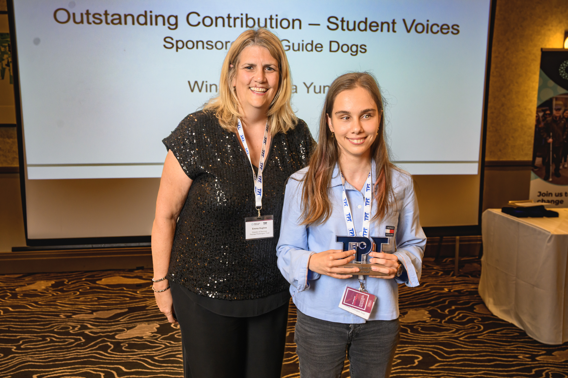 Zehra, Student Voices volunteer, stands with Director of Services, Emma Huges at the 2026 Rodney Powell Awards. Zehra is holding a TPT shaped trophy as the winner of the Outstanding Contribution to Student Voices award. A screen behind them displays the text “Outstanding Contribution – Student Voices,” sponsored by Guide Dogs, and identifies the winner as Zehra Yunel. 