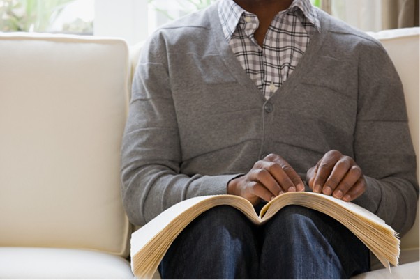 An older man is sat on a sofa reading braille from a book.