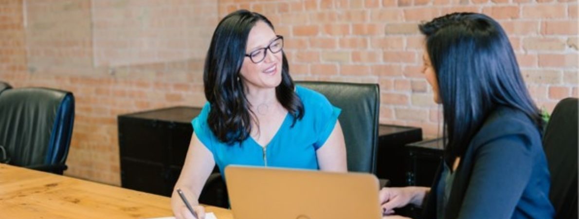 Two women are sat on the same side of a wooden table facing each other in conversation. One woman whose face can be seen is smiling, wearing glasses, and taking notes on an A4 paper notepad. The other woman whose face cannot be seen has a laptop computer on the table in front of her. The laptop is angled so both women can see the screen. The second women is not using the laptop as her attention is focused on the first woman and their conversation.