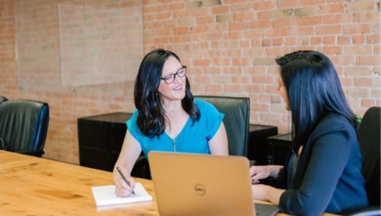 Two women are sat on the same side of a wooden table facing each other in conversation. One woman whose face can be seen is smiling, wearing glasses, and taking notes on an A4 paper notepad. The other woman whose face cannot be seen has a laptop computer on the table in front of her. The laptop is angled so both women can see the screen. The second women is not using the laptop as her attention is focused on the first woman and their conversation. | links to article: Is College for Me? A Guide for Blind and Partially Sighted Adult Learners