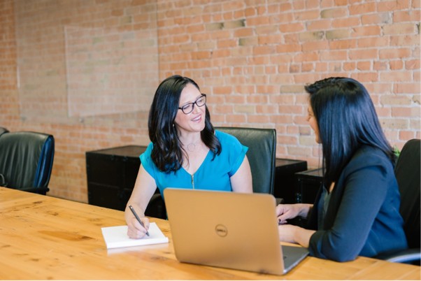 Two women are sat on the same side of a wooden table facing each other in conversation. One woman whose face can be seen is smiling, wearing glasses, and taking notes on an A4 paper notepad. The other woman whose face cannot be seen has a laptop computer on the table in front of her. The laptop is angled so both women can see the screen. The second women is not using the laptop as her attention is focused on the first woman and their conversation.