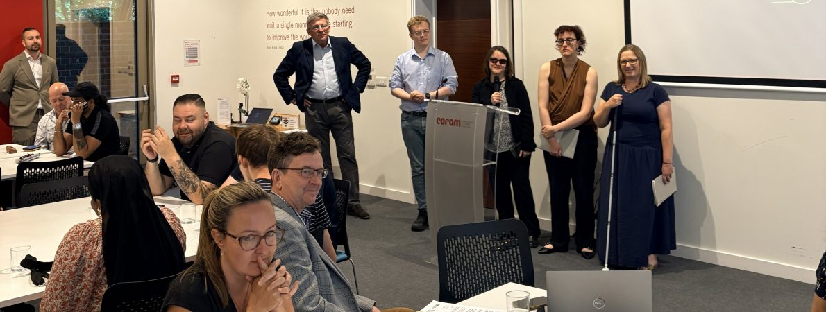 Several people presenting at the front of a conference room while attendees sit at tables and listen during a workshop or meeting.