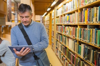 An older man is in a library stood parallel to a bookshelf extending behind him. He is holding a tablet he is looking at to search for information. He wears a book bag over his shoulder. Another student can be seen just out of frame at a desk head down studying.