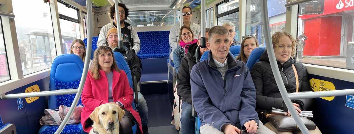 Photo of a group of people sitting on a bus. They are all smiling. There is a guide dog in a harness sitting on the floor in front of one of the people.