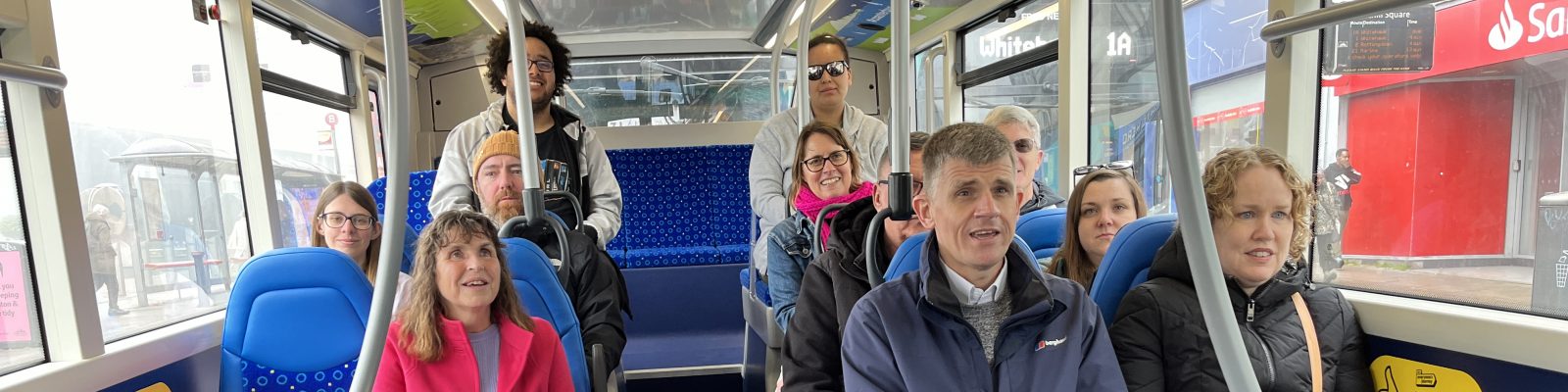 Photo of a group of people sitting on a bus. They are all smiling. There is a guide dog in a harness sitting on the floor in front of one of the people.