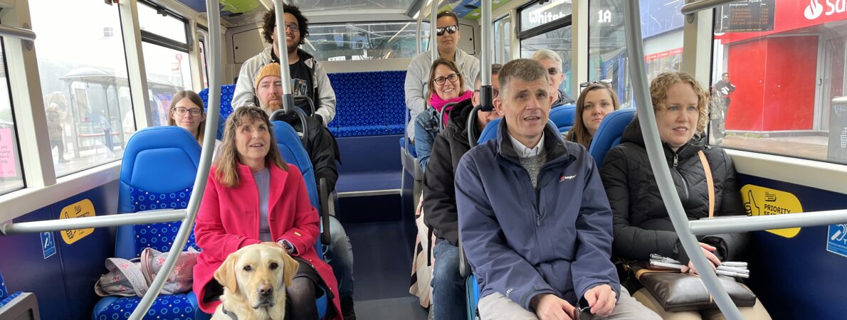 Photo of a group of people sitting on a bus. They are all smiling. There is a guide dog in a harness sitting on the floor in front of one of the people.