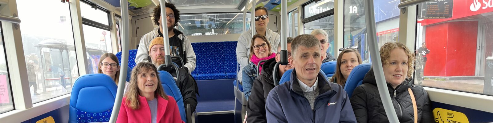 Photo of a group of people sitting on a bus. They are all smiling. There is a guide dog in a harness sitting on the floor in front of one of the people.