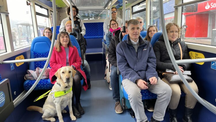 Photo of a group of people sitting on a bus. They are all smiling. There is a guide dog in a harness sitting on the floor in front of one of the people. | links to article: Catch the Bus Month: Helping every blind and partially sighted passenger feel seen, supported and confident