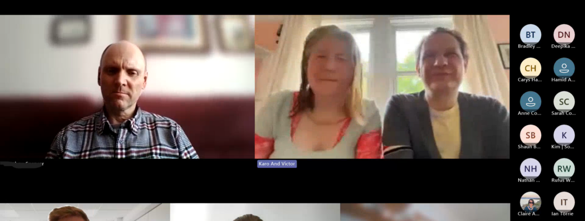 Screenshot of a virtual meeting in a video conferencing platform displayed in gallery view. Five participants have live video feeds enabled, arranged in a grid across the screen. Two larger video windows appear along the top row and three smaller video windows appear along the bottom row. Several additional participants are represented by circular profile icons with initials in a vertical participant panel on the right-hand side of the screen. The top-left video tile shows a person wearing a check-pattern shirt, seated indoors against a blurred background with framed pictures on a wall. The top-center video tile shows two people sitting side by side in front of a bright window with curtains, creating strong backlighting. A label beneath this video feed reads “Karo And Victor.” The bottom row contains three video feeds. The left feed shows a person wearing glasses seated in an office environment. A standing banner with the letters “TPT” is visible in the background. The centre feed shows a person seated against a plain light-coloured wall, resting clasped hands near their face. The right feed shows a person in profile seated indoors, with a blurred shelving unit visible behind them.