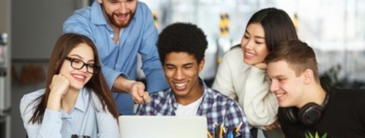 A group of students working around a laptop, sitting at a table covered with open books, pens and paper, getting stuck into their work.
