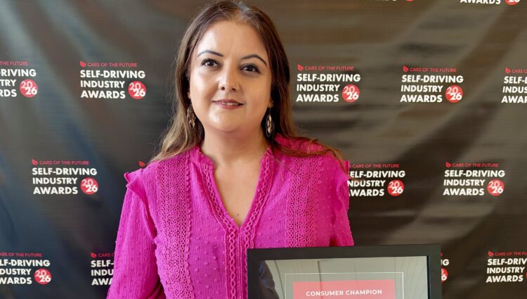 Bhavini, who is wearing a pink blouse and smiling, is standing in front of a step and repeat board for the Self-Driving Industry Awards 2026. She is holding a framed award certificate, as she has won the Consumer Champion Award 2026.